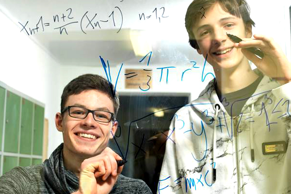 Zwei Schüler stehen hinter einer Glastür und schreiben mit Filzstiften mathematische Gleichungen auf die Scheibe.