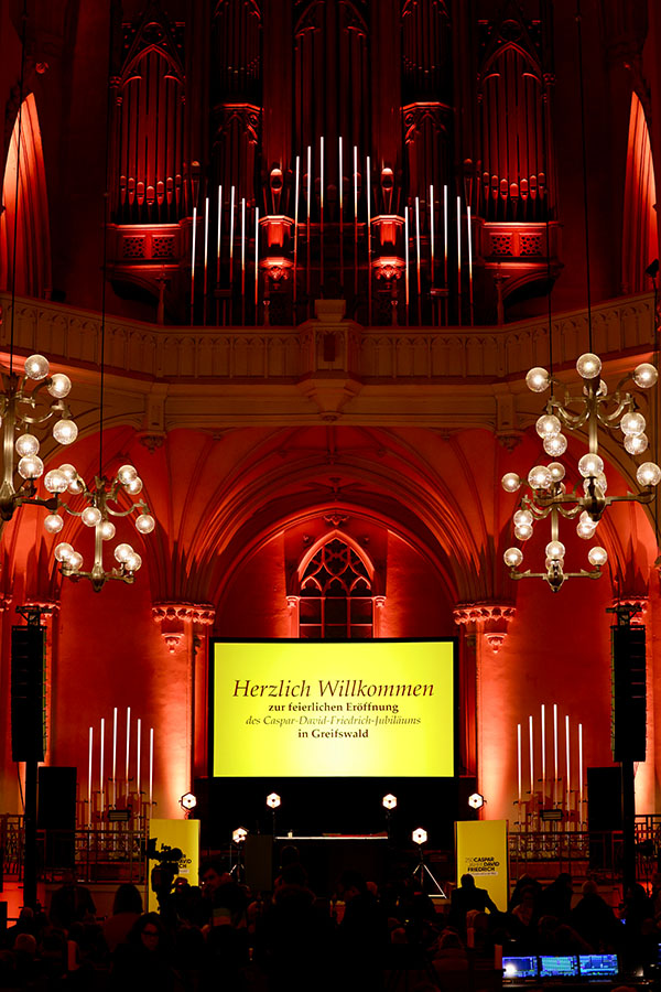 Auf dem Foto steht auf einem Display mit gelbem Hintergrund, umgeben von rot angestrahlten Wänden im Dom "Herzlich willkommen zur feierlichen Eröffnung des Caspar-David-Friedrich-Jubiläums in Greifswald"